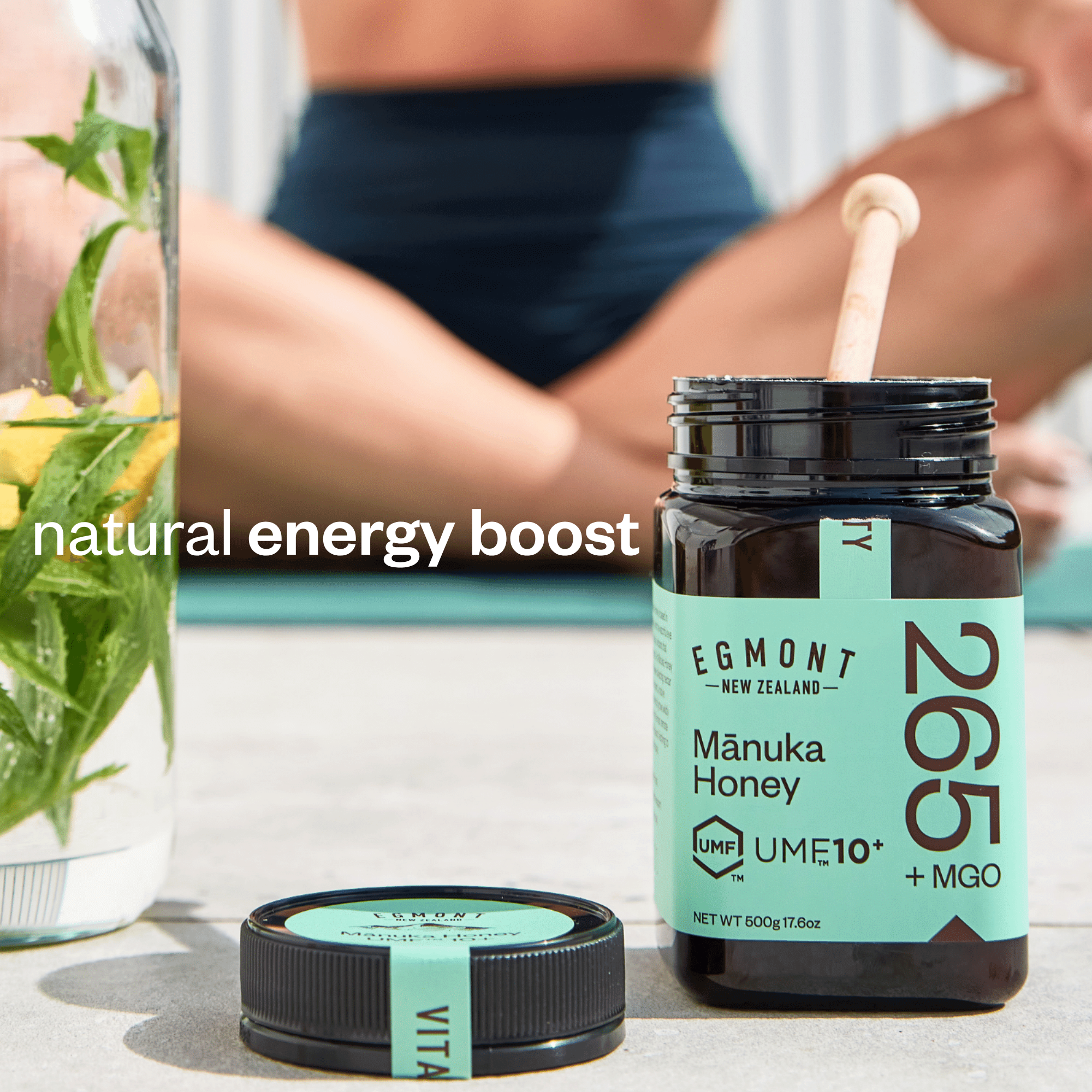 An open jar of Egmont New Zealand Mánuka Honey with a wooden dipper, shown alongside its cap on a light stone surface. A blurred person in swim trunks sits in the background, and a glass of mint water is visible on the left.