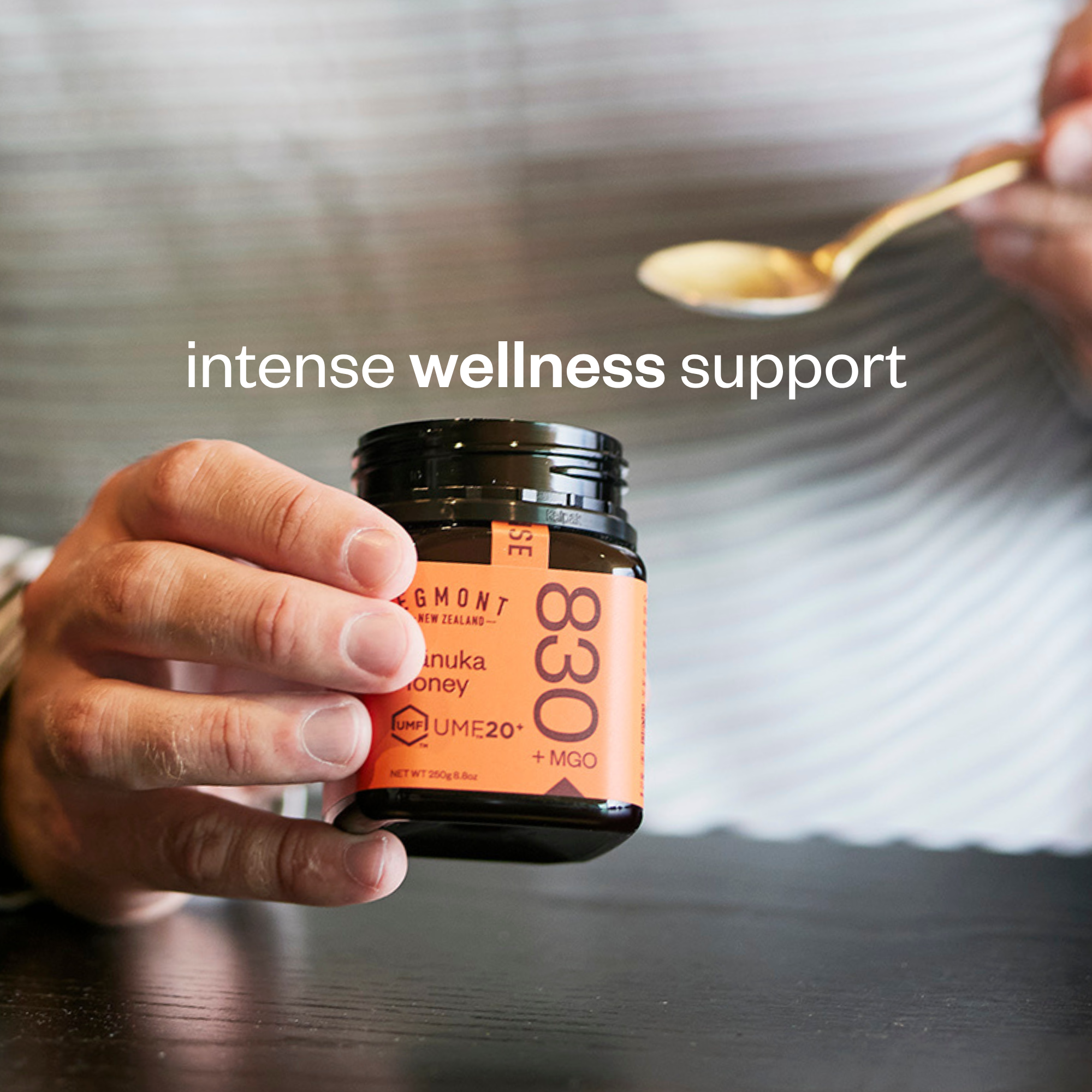 A hand holds a small jar of UME20+ Manuka Honey with an orange label, while another hand holds a spoon above it. White text overlay reads 'intense wellness support'.